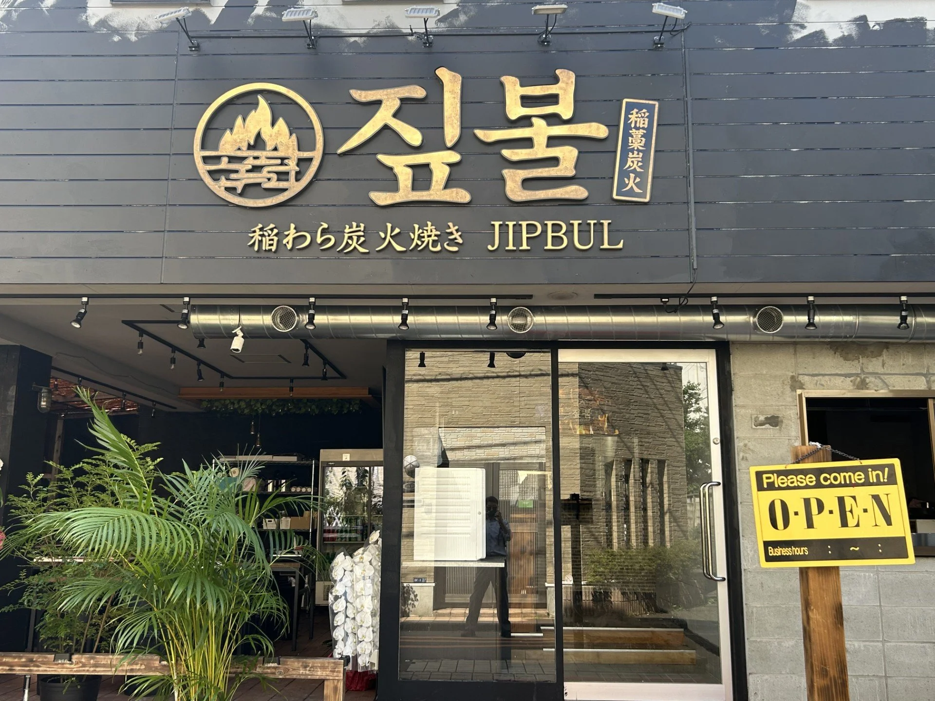 炭火の焼き網で肉が大きく焼け、奥に笑顔で食事を囲む卓が見える店内の場面